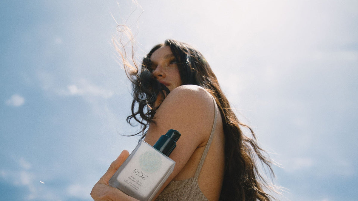 A woman with long, loose dark hair holds a bottle of RŌZ Milk Hair Serum against her shoulder under a blue sky.