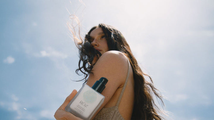 A woman with long, loose dark hair holds a bottle of RŌZ Milk Hair Serum against her shoulder under a blue sky.